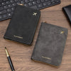A black passport wallet with gold stitching and a small airplane emblem is shown next to a laptop keyboard and a pen on a woo