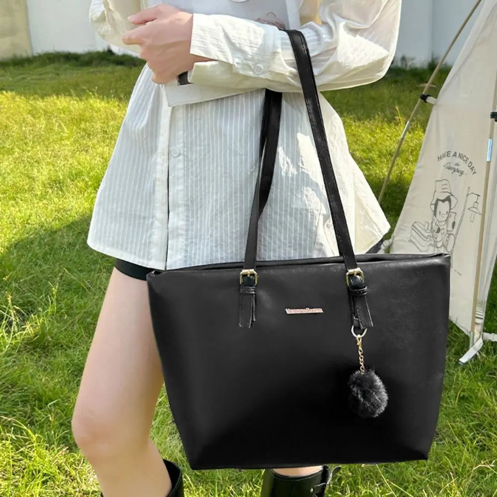 Una bolsa tote de cuero sintético negro con un dije dorado y un pompón negro sostenida por una persona al aire libre.