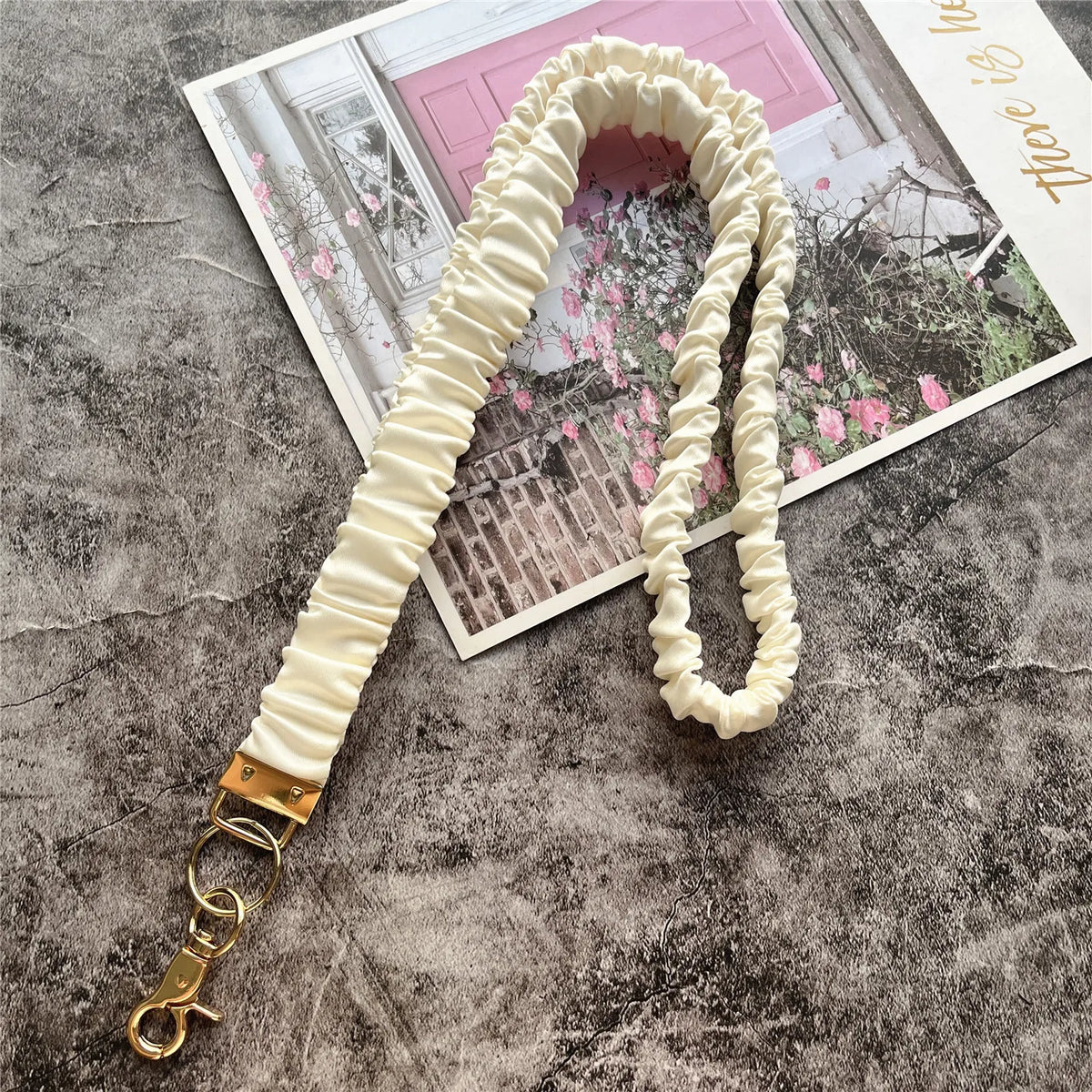 Cremfarbener, geraffter Lanyard mit goldenem Schlüsselclip auf grauer Oberfläche neben einem Foto.