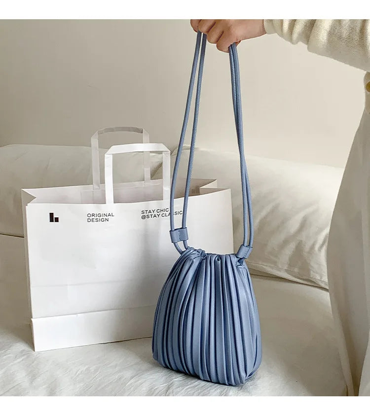 A light blue PU leather pleated crossbody bag is being held by a hand, placed in front of white paper shopping bags.