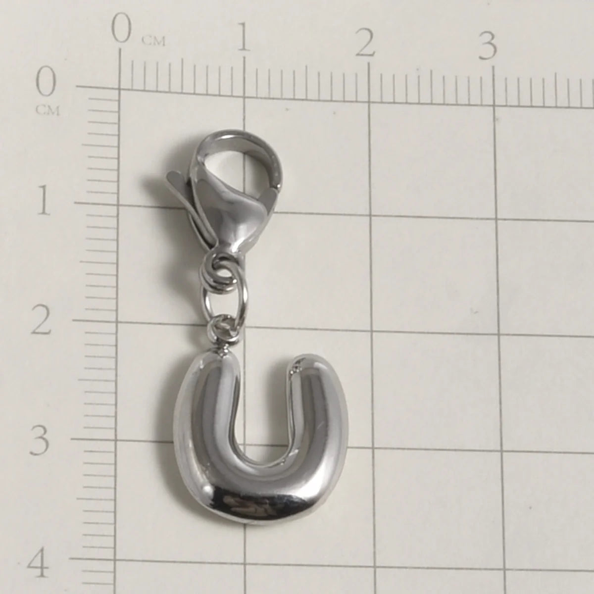 A silver stainless steel alphabet letter charm shaped like the letter U, displayed on a ruler.