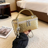 A close-up shot of a gold woven leather crossbody bag with a chain strap being held by a hand on a light carpet.
