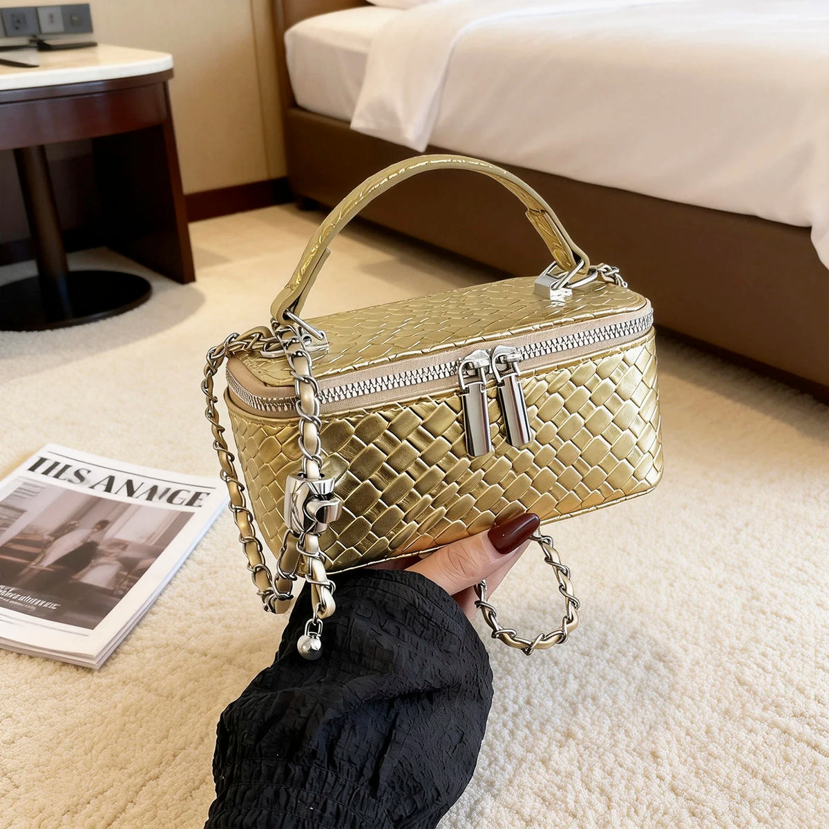 A close-up shot of a gold woven leather crossbody bag with a chain strap being held by a hand on a light carpet.
