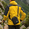 A man wearing a yellow and black jacket with a black sling bag on his back, hiking in a mountainous outdoor setting.
