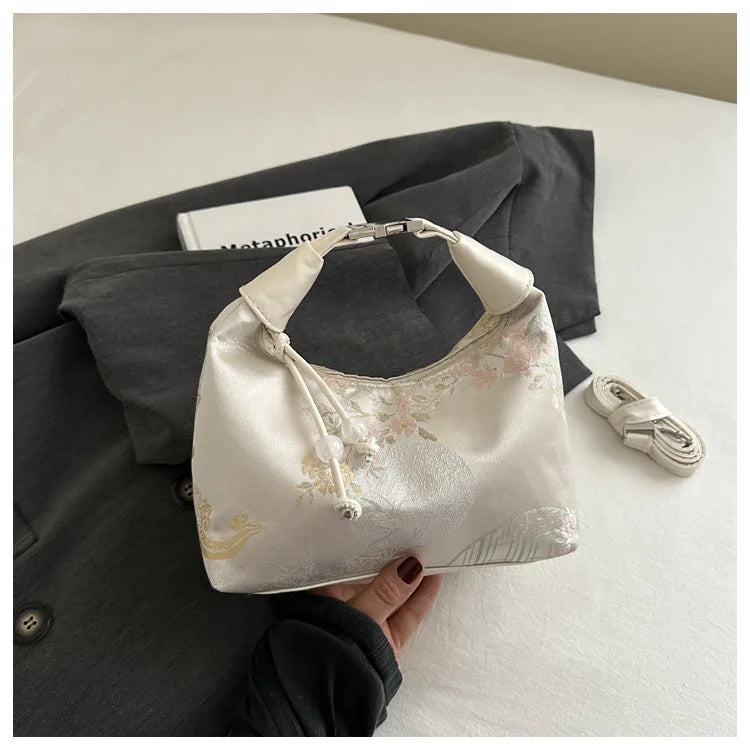 A cream silk embroidered crossbody shoulder bag is shown resting on a dark gray blazer.