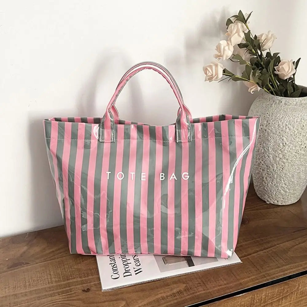 A striped transparent PVC tote bag with pink and green vertical stripes resting on a wooden surface.