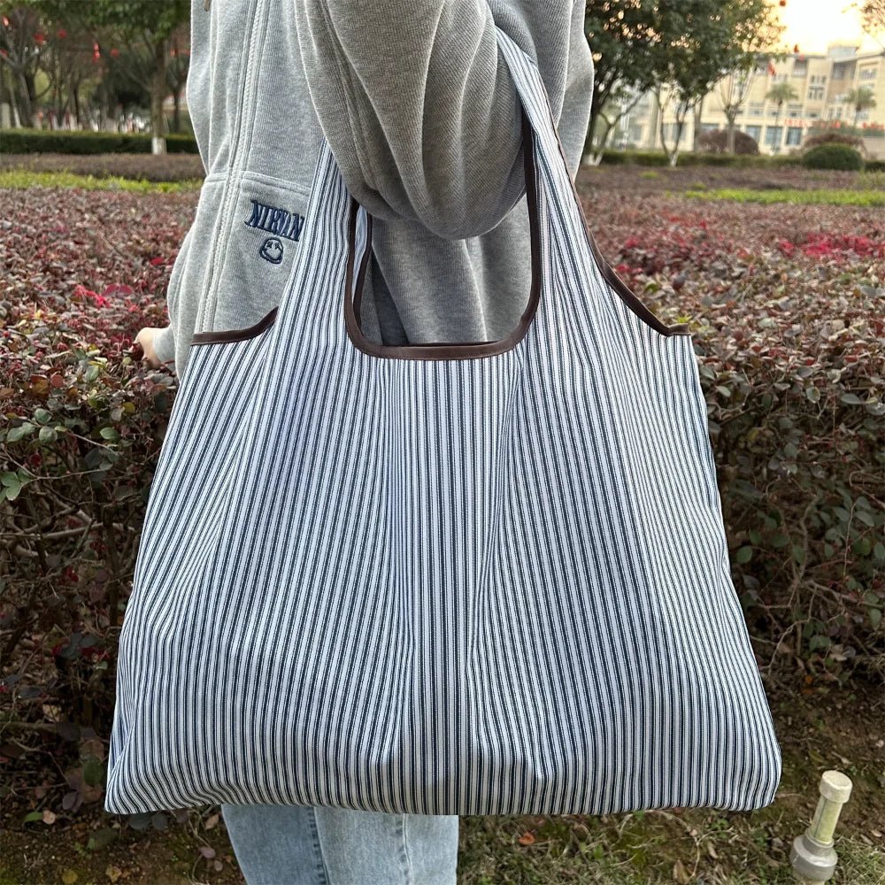 A person holds a striped grocery tote bag with a brown handle against a blurred outdoor background.