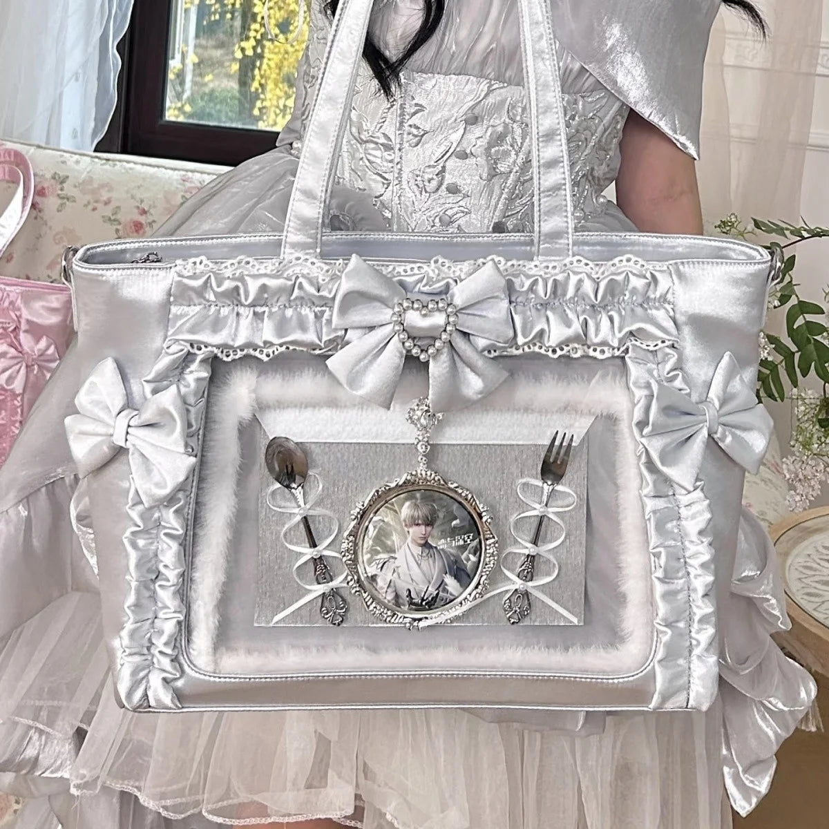 A silver and white transparent shoulder tote bag with decorative bows and a lace trim carried by a person wearing a light-col