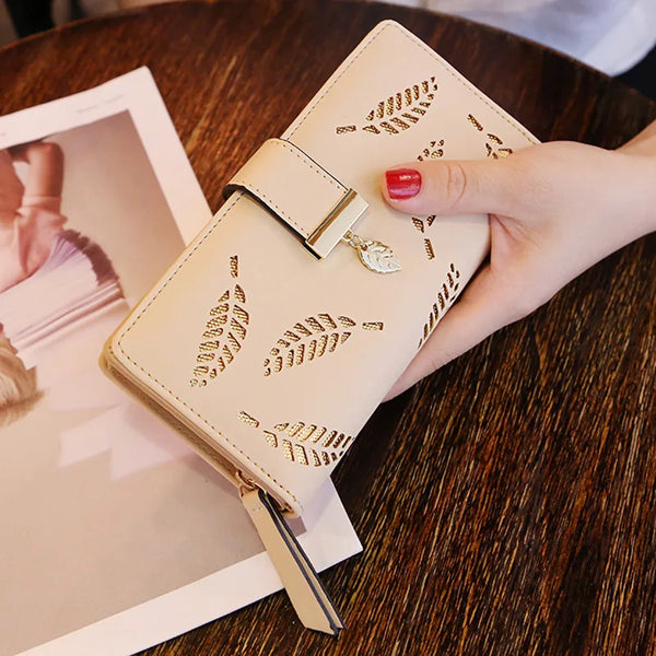 A hand holds a beige PU leather statement clutch wallet with gold embossed leaf patterns on a dark wooden table.