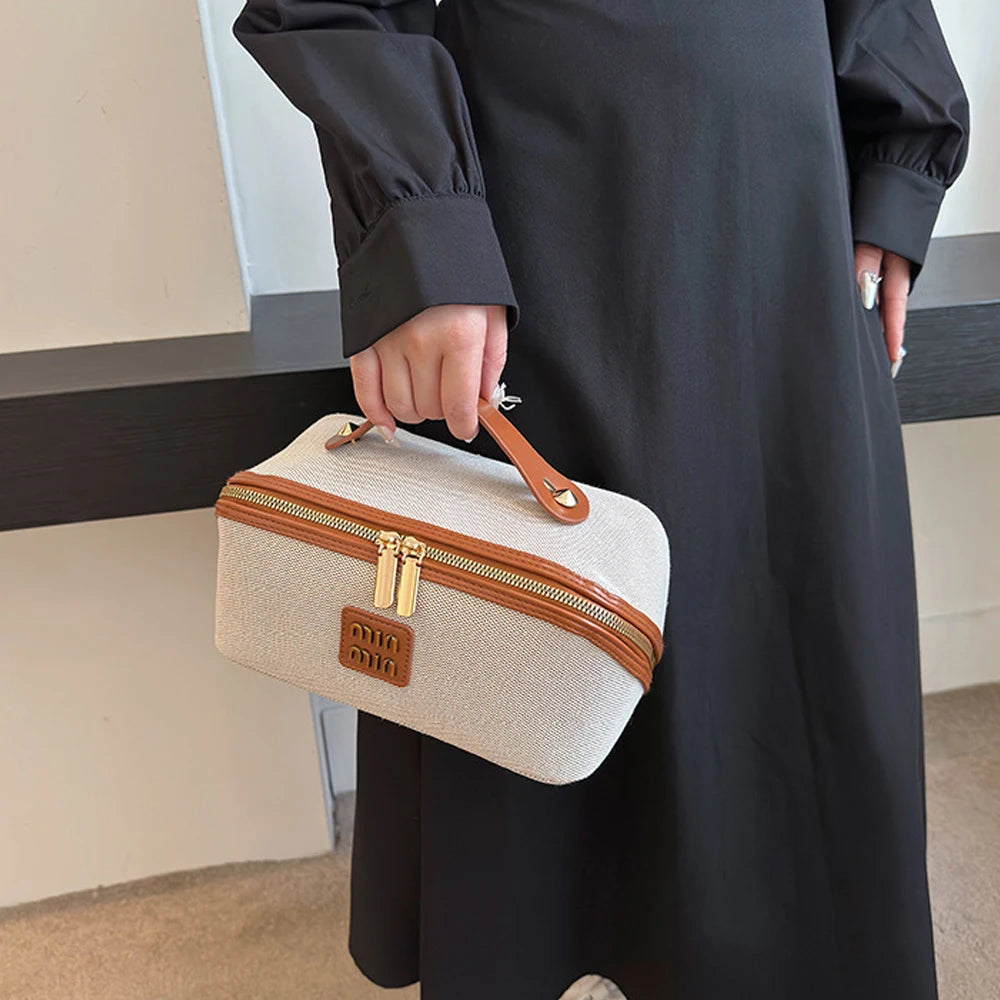 A person holds a beige travel toiletry bag with a brown leather strap and gold zippers against a dark dress.