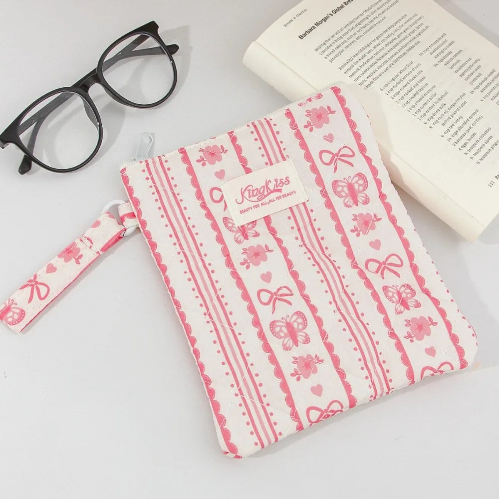 A pink and white floral plaid cotton tablet pouch with a ribbon closure next to eyeglasses and a book.