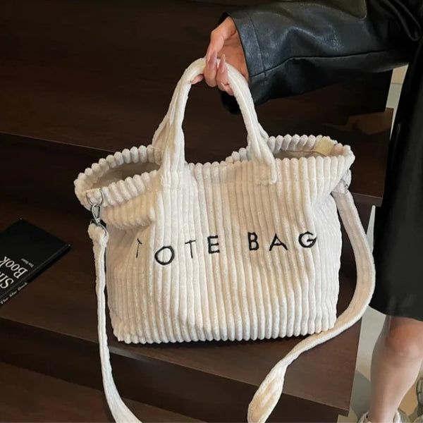 Eine cremefarbene, gestreifte Samt-Schultertasche wird von einer Person vor einer dunklen Holzoberfläche gehalten.
