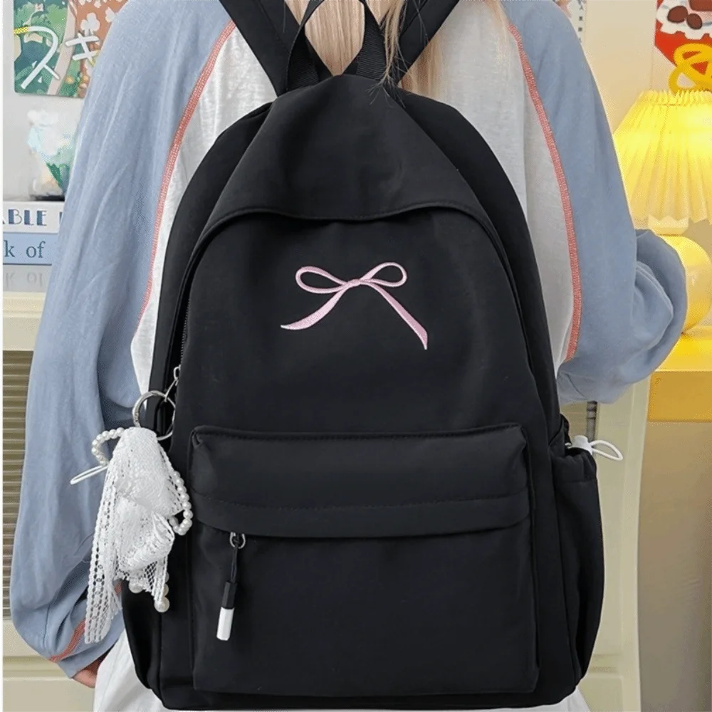 A black backpack with a pink bow embroidery and a white tassel attached to the zipper.