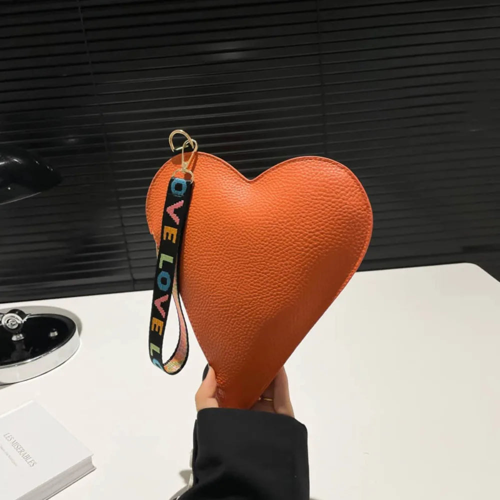Cartera de monedas con forma de corazón naranja de PU y correa de cuero negro, sostenida contra un fondo oscuro.