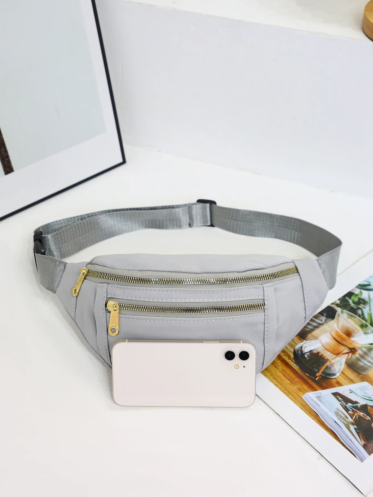 A light gray nylon crossbody fanny pack with a zippered pocket and a smartphone resting on it.