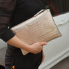A person holds a gold-toned alligator print clutch bag against a white car door.