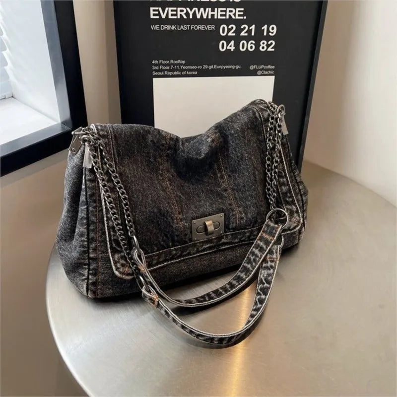 A dark gray denim tote bag with a textured finish and a silver chain strap is displayed on a light surface.