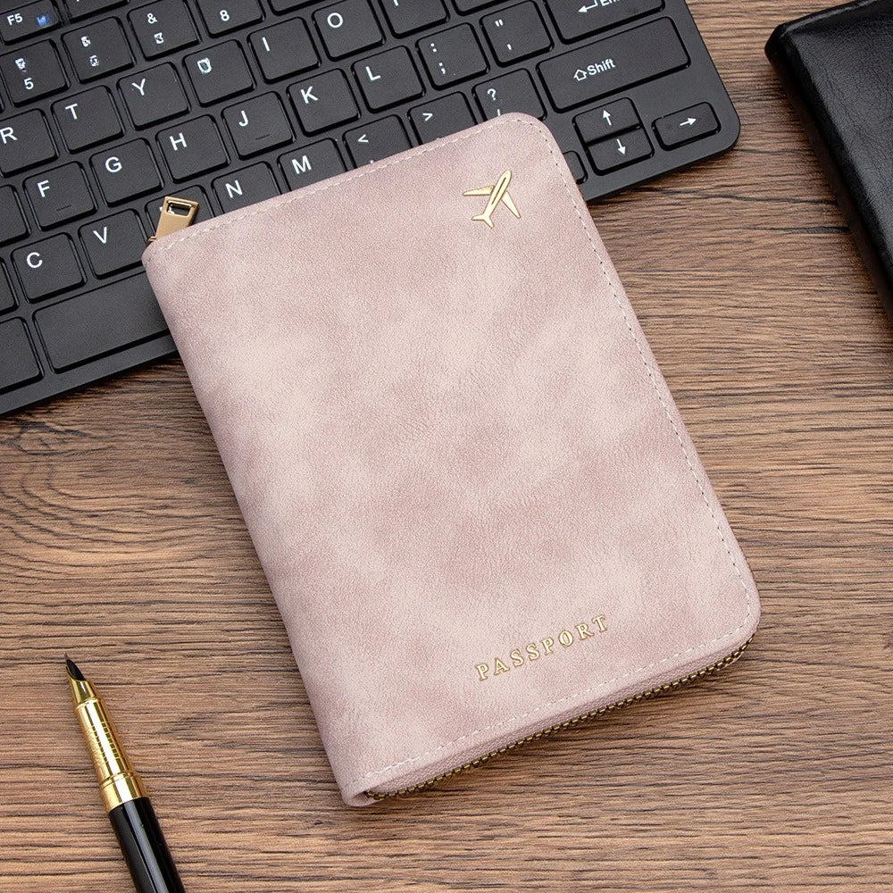 A light pink slim passport wallet with a gold embossed airplane logo resting on a wooden desk next to a laptop keyboard.