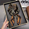 A dark patterned silk hair scarf tied in a bow is displayed in a black frame on a gray surface.