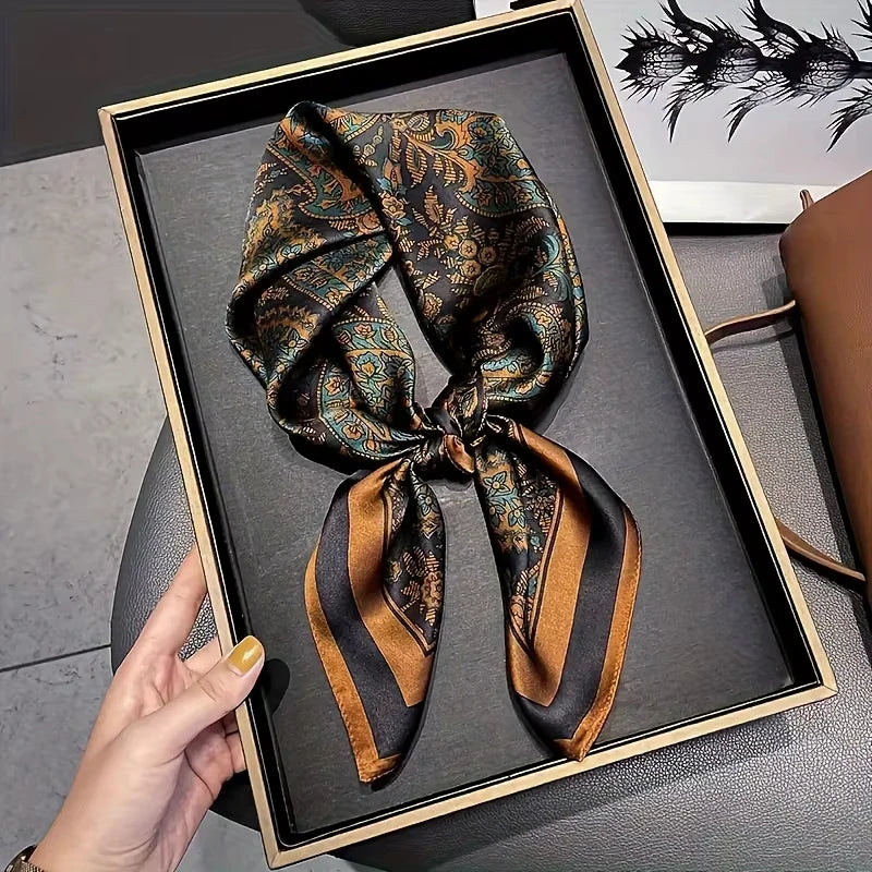 A dark patterned silk hair scarf tied in a bow is displayed in a black frame on a gray surface.