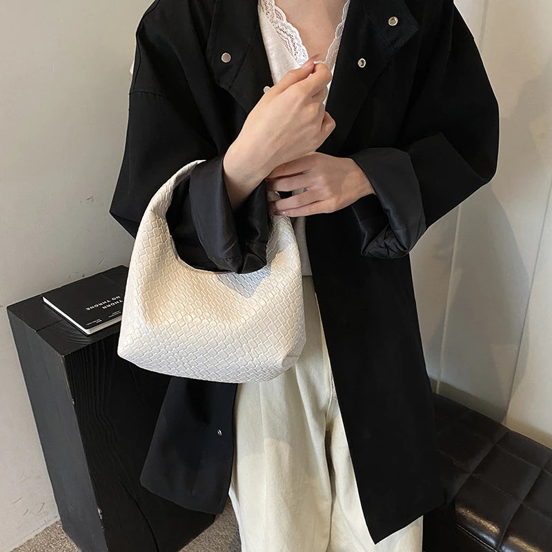 A woman holds a woven embossed hobo shoulder bag with a top handle in front of a black coat and light-colored pants.