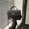 A woman holds a black textured shoulder messenger bag with a gold butterfly charm against a light background.