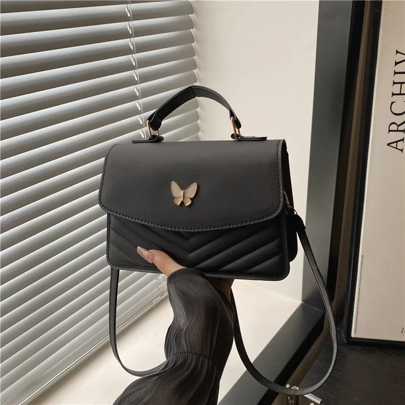 A woman holds a black textured shoulder messenger bag with a gold butterfly charm against a light background.