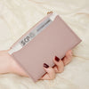 Une main tient un porte-cartes en cuir PU rose pâle minimaliste avec une boucle dorée sur fond crème.