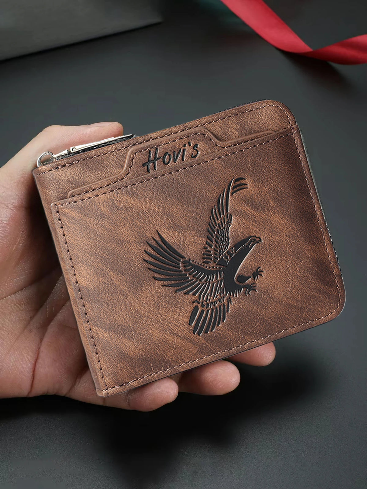 A brown embossed PU leather slim wallet with a black eagle design is held in a hand.