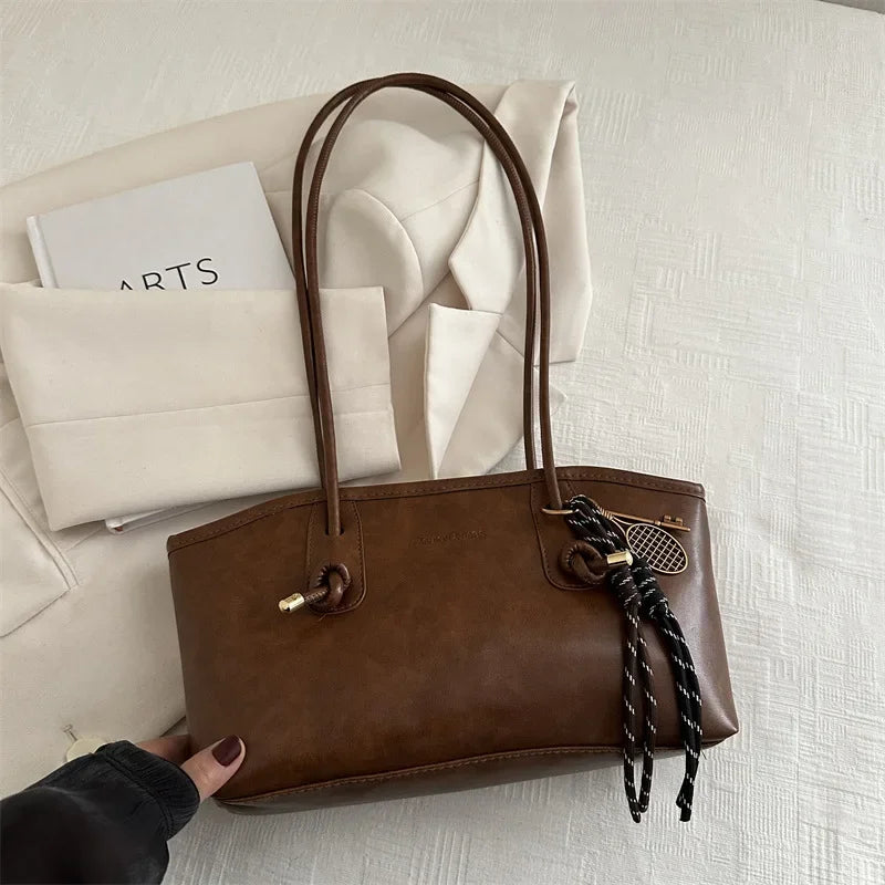 A brown leather tote bag with a handle and decorative chain is placed on a white textured surface next to a notebook.