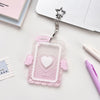 A clear pink acrylic photocard holder with a heart design and silver charm attached to a keyring.