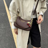 A woman wearing a beige trench coat and black skirt holds a brown leather shoulder bag with gold chain details.