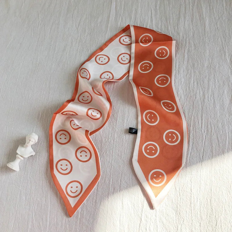 Écharpe de soie orange avec des visages souriants blancs drapée sur une surface blanche.