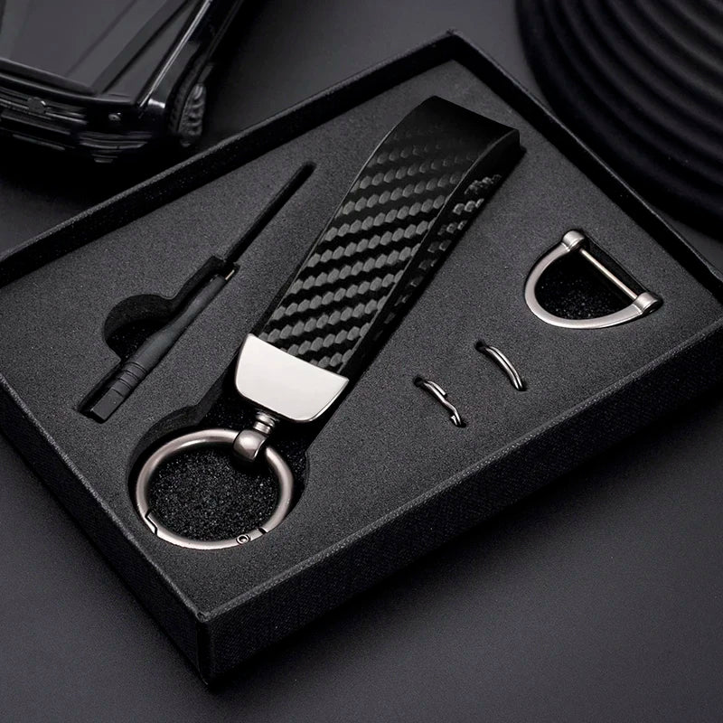 A black box holds a carbon fiber leather keychain with a silver clasp and a small tool.
