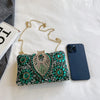 A sparkling green and gold rhinestone evening clutch bag resting on a white surface next to a smartphone.