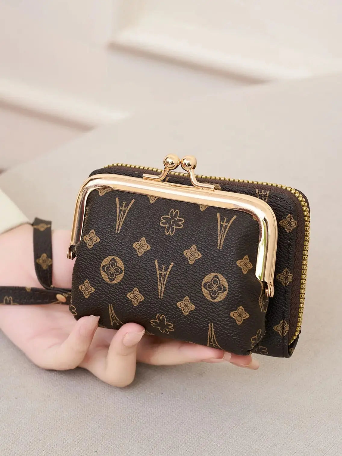 A dark brown fashion print accordion card holder wallet with gold-toned hardware and a black strap is held in a hand.