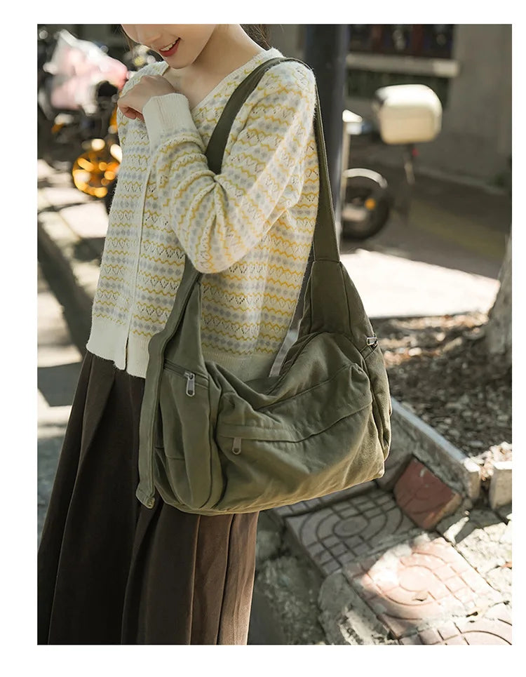 A woman wearing a yellow and white patterned sweater holds a large olive green cotton canvas shoulder crossbody bag.