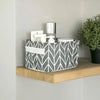 A gray and white patterned storage basket filled with white bathroom or desk organization items on a wooden shelf.