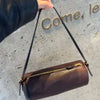 A dark brown vintage cow leather cylinder shoulder bag with a gold zipper and black strap is held against a textured gray wal