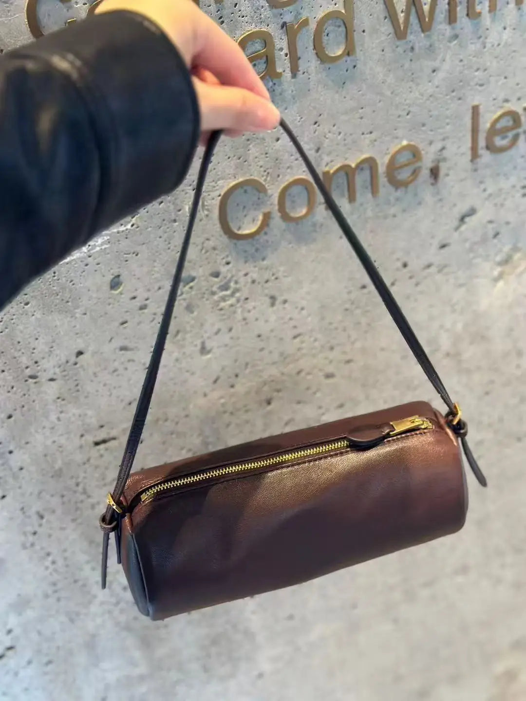 A dark brown vintage cow leather cylinder shoulder bag with a gold zipper and black strap is held against a textured gray wal
