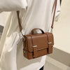 A brown minimalist leather crossbody bag with a leather strap is shown against a white shirt.