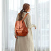 A woman wearing a tan coat is standing indoors, carrying a brown vintage soft leather backpack.