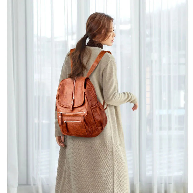 A woman wearing a tan coat is standing indoors, carrying a brown vintage soft leather backpack.
