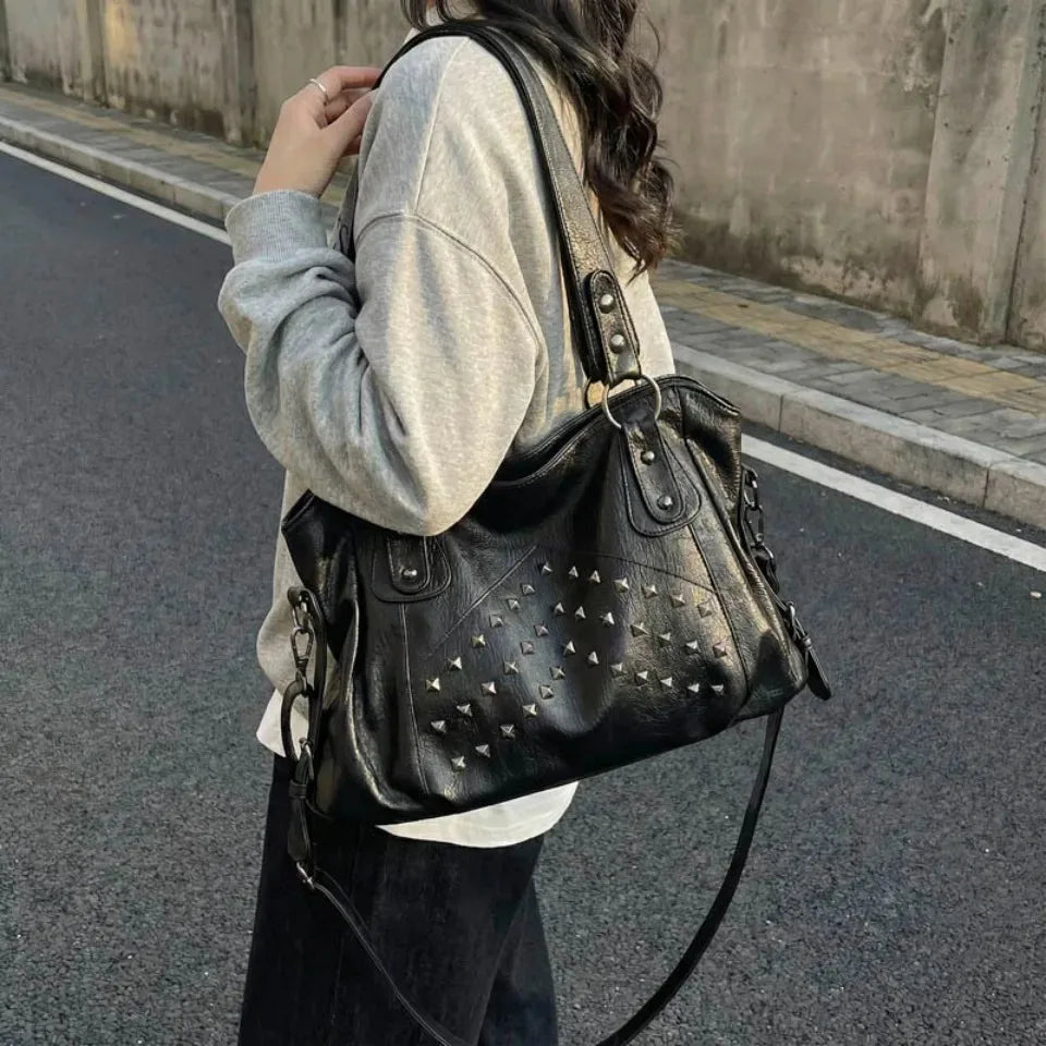 A woman is holding a black leather tote bag with silver rivets and a strap, standing on an asphalt road.