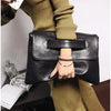 A hand holds a black leather clutch bag with a silver chain bracelet against a beige sweater.