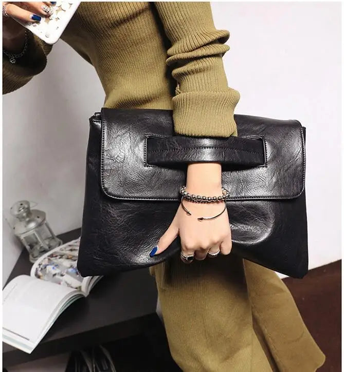 A hand holds a black leather clutch bag with a silver chain bracelet against a beige sweater.