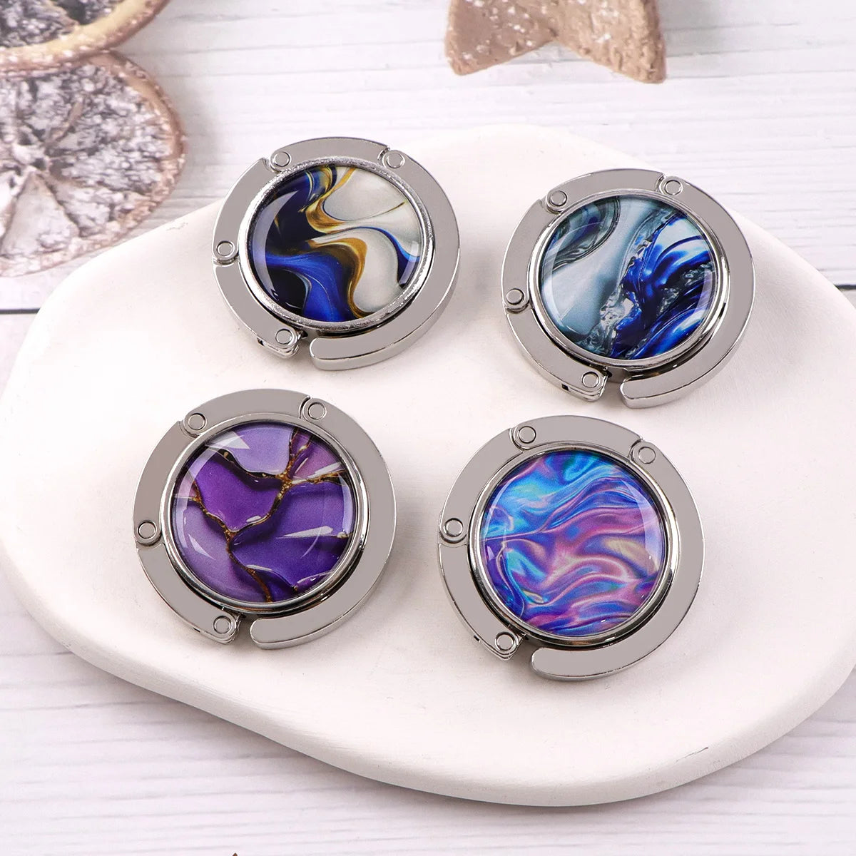 Four silver folding table bag hooks with marbled blue and purple gemstone designs on a white plate