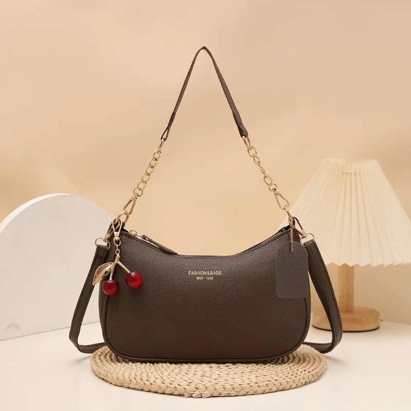 A dark brown leather shoulder bag with a gold chain and cherry pendant resting on a woven mat.