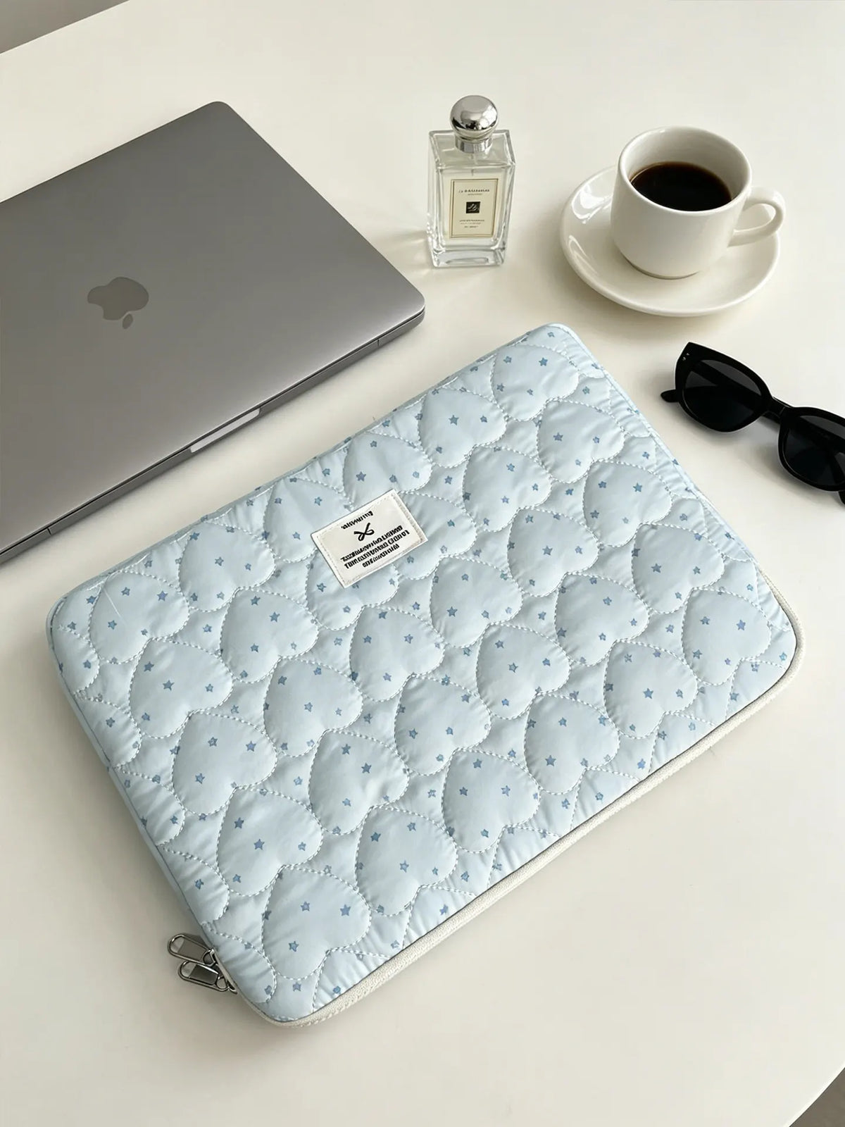 Light blue quilted laptop sleeve with a zipper and small silver perfume bottle on a white table.