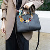 A woman holds a black leather handbag with a multi-color acrylic chain strap, paired with a beige coat and black pants.